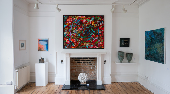 Gallery space with brightly coloured painting over a fire place with sculptures on either side