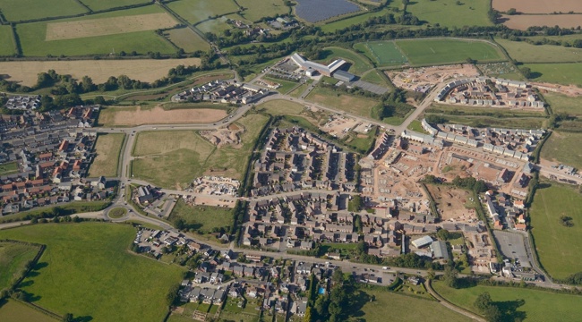 An aerial photograph of Cranbrook Town Centre with the pub and roundabout to the south, education campus to the north and housing east and west. The town centre is the square area of open land left of centre.