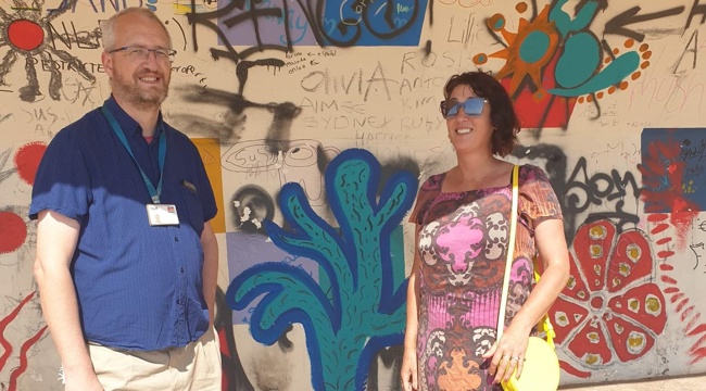 Man in blue on the left next to woman in pink top and sunglasses standing in front of brightly coloured mural