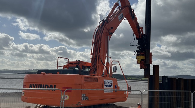A construction vehicle installing a steel sheet pile wall on Exmouth seafront. The sea and sky are in the background