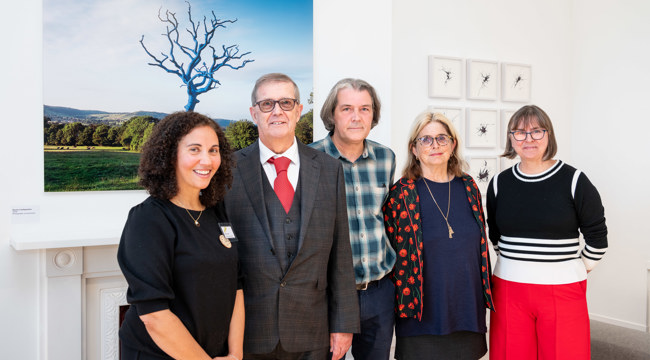 Group of 3 women and 2 men in a white gallery space with photograph on the wall behind them
