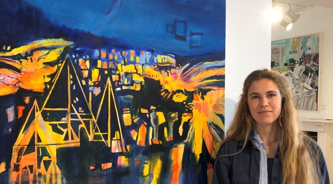 Girl with long blonde hair stands next to an abstract painting of blue and yellow night scene 