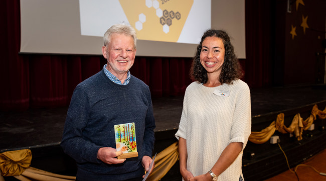 Man with grey hair in navy top is smiling and holding glass award next to a woman with black hair and white top